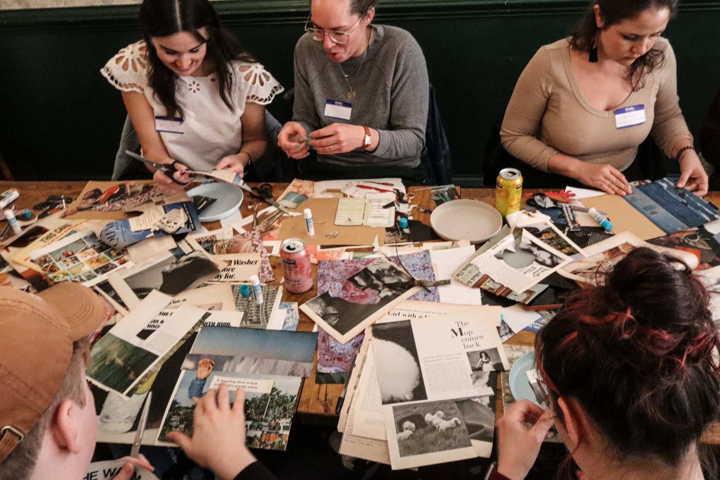 People sitting at a table covered with magazines, newspapers, and craft supplies, engaged in a creative activity.
