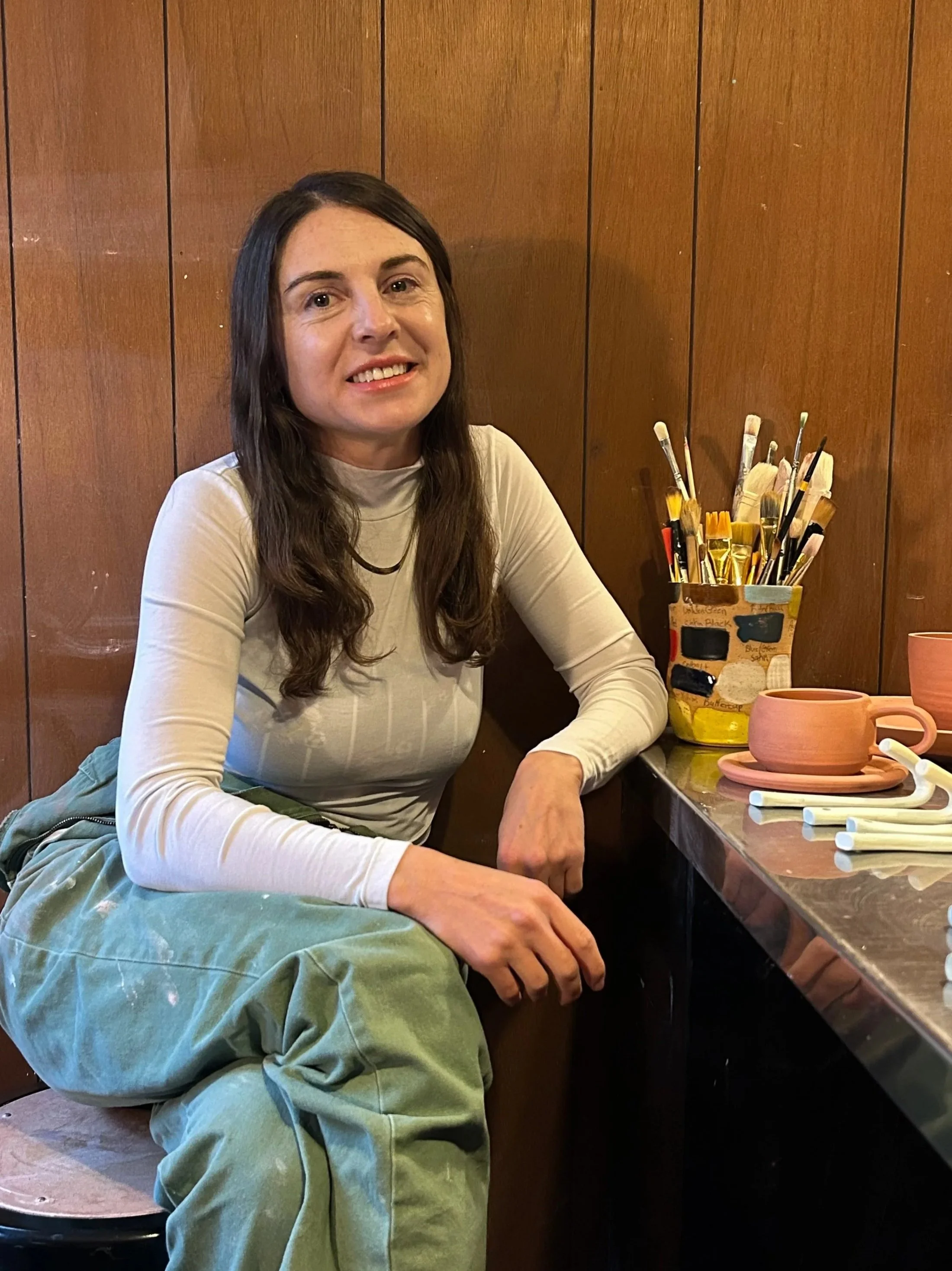 A woman with long dark hair and a light-colored long sleeve shirt, sitting at a table in front of a wooden-paneled wall, with various paintbrushes and pottery on the table.