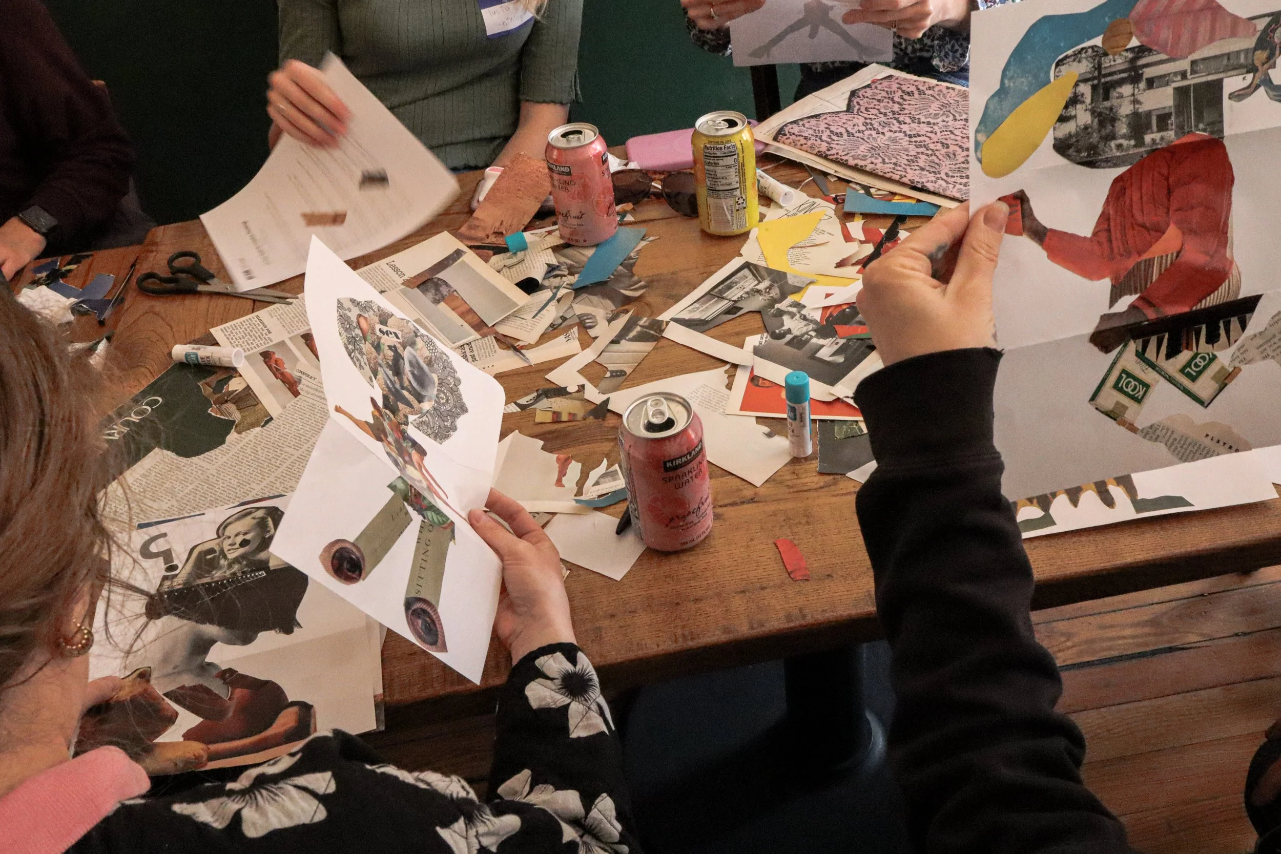People gathered around a wooden table working on collage art projects with scissors, glue sticks, magazines, and printed images, including cartoons and magazine clippings.