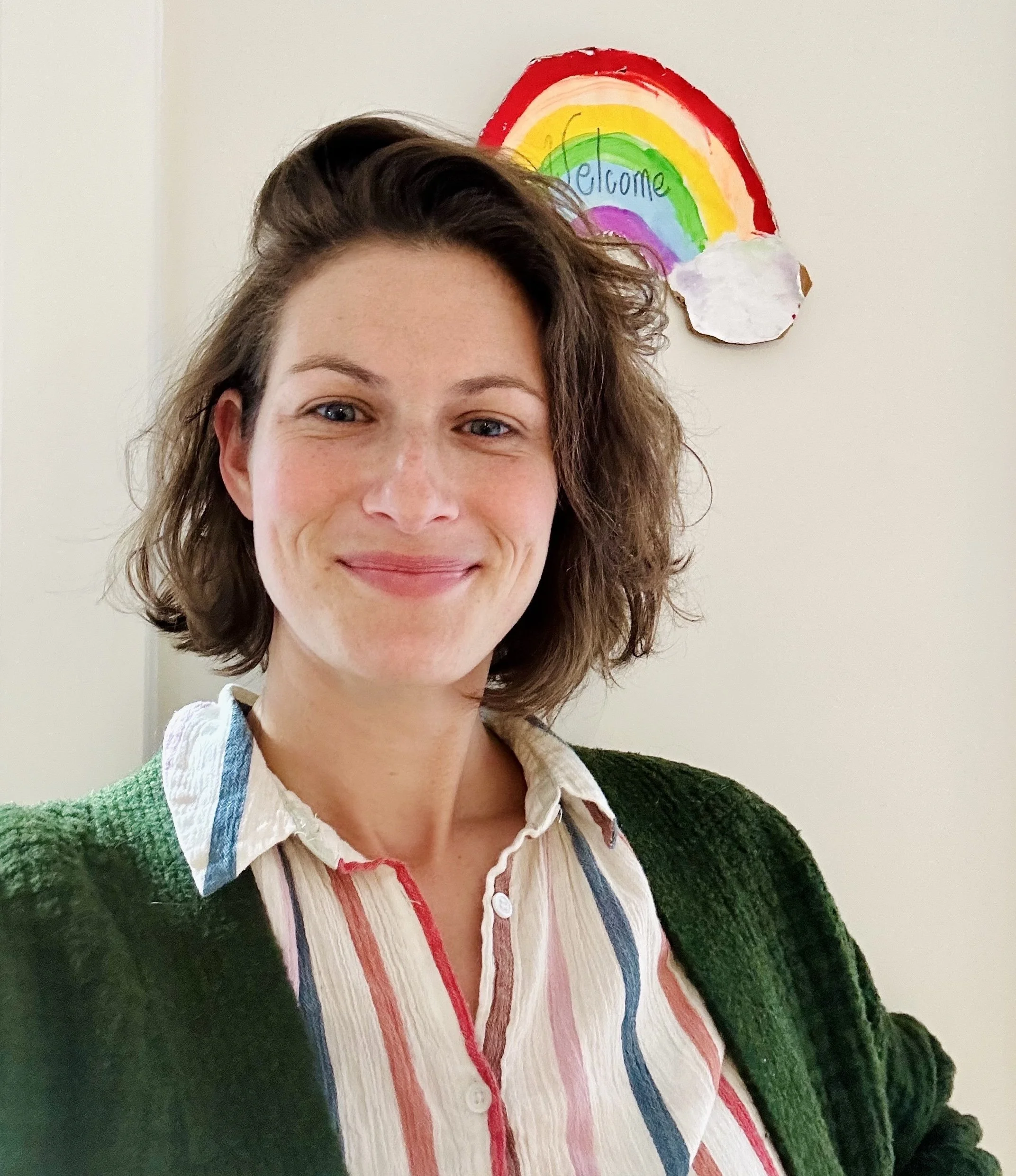 Close-up of a smiling woman with short wavy hair wearing a colorful striped shirt and a green cardigan, standing in front of a wall with a rainbow drawing containing the word 'welcome'.