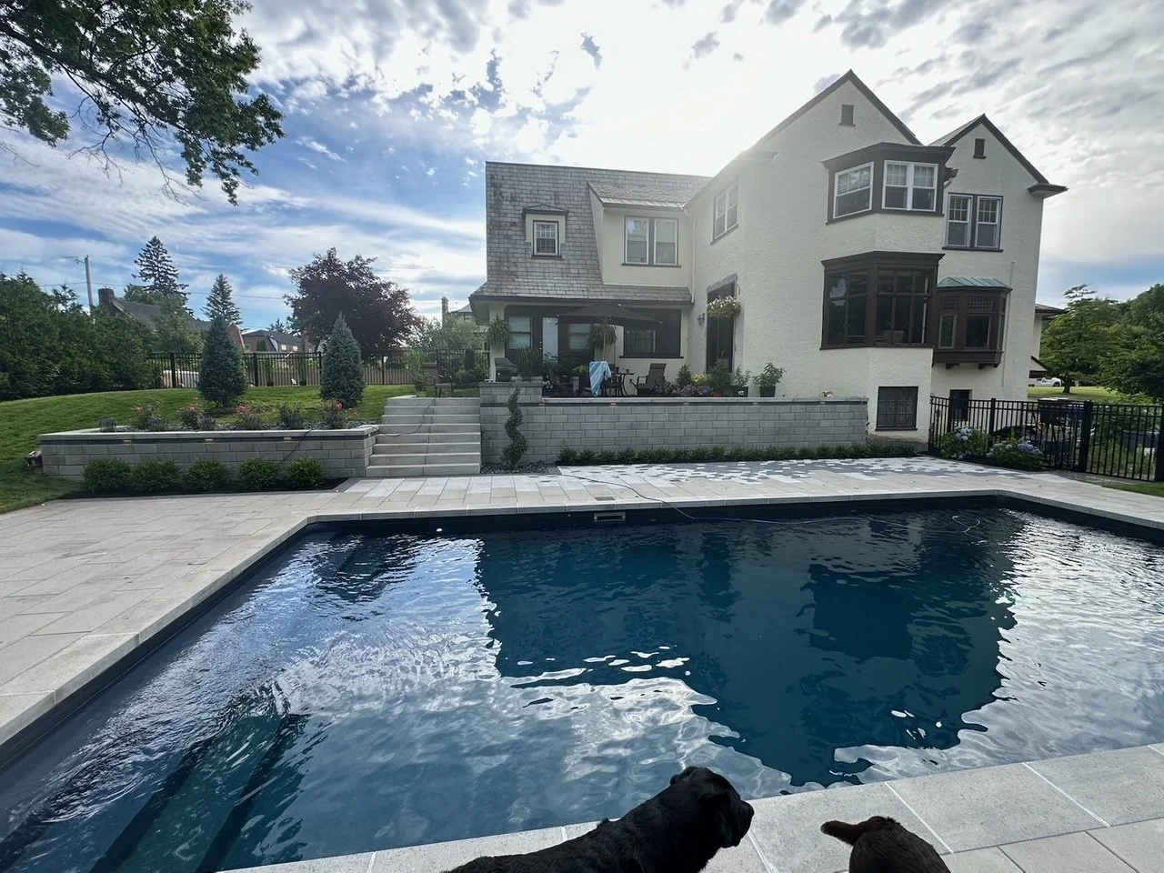 View of a large backyard with a swimming pool, patio, and a white house with multiple levels and black window frames, under a partly cloudy sky.