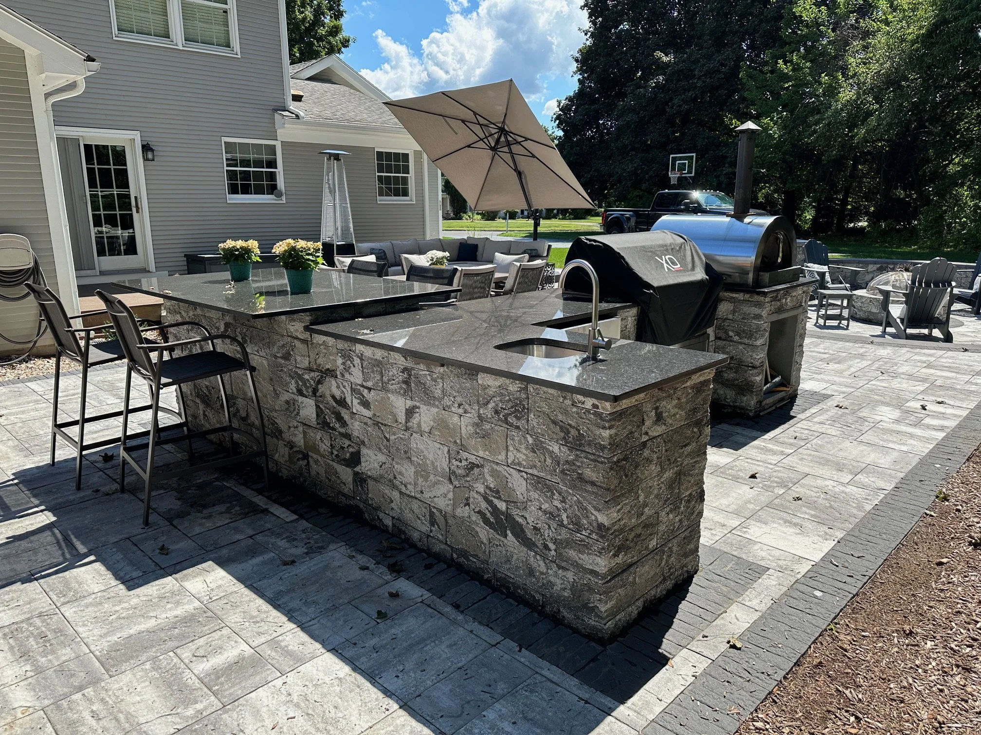 An outdoor patio with a section of a house in the background. The patio features a stone bar with a granite countertop, barstools, and a built-in grill with a cover. There are also outdoor seating arrangements including a table with potted flowers and a sitting area with an umbrella. The area is paved with stone tiles and has a natural grassy area with a basketball hoop and a truck in the background.