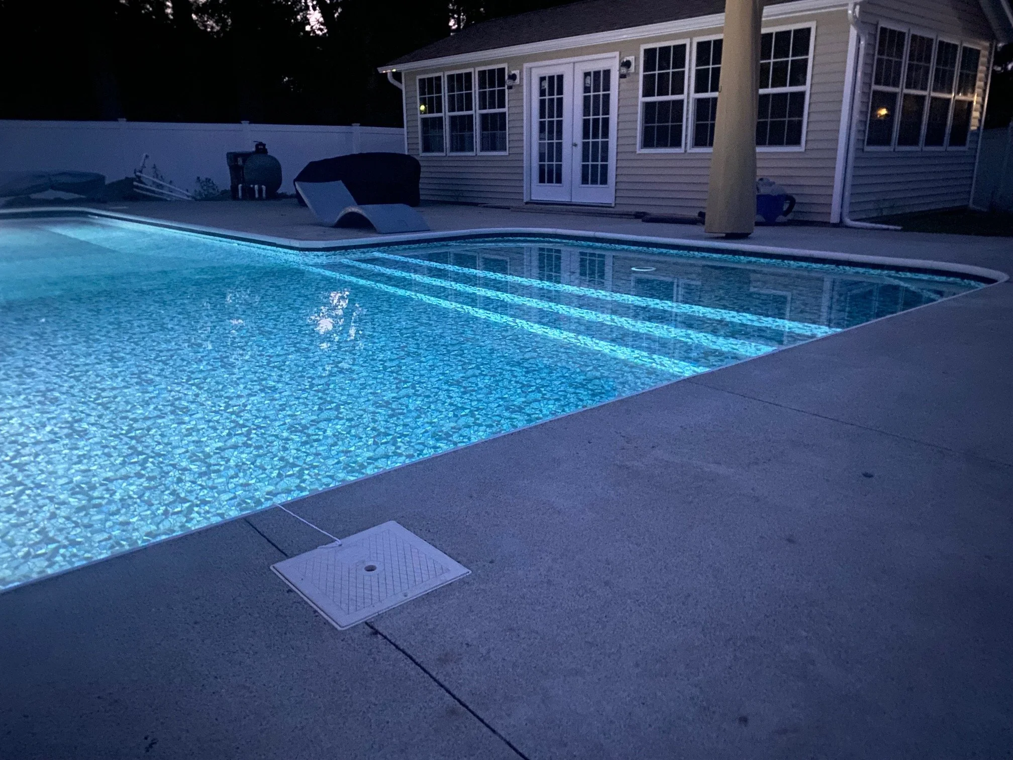 Backyard swimming pool with illuminated water and pool steps at night. A house with windows and a deck are visible in the background.