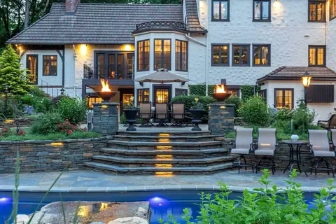 Backyard with a pool, stone patio, and outdoor seating area, with a large white house in the background.
