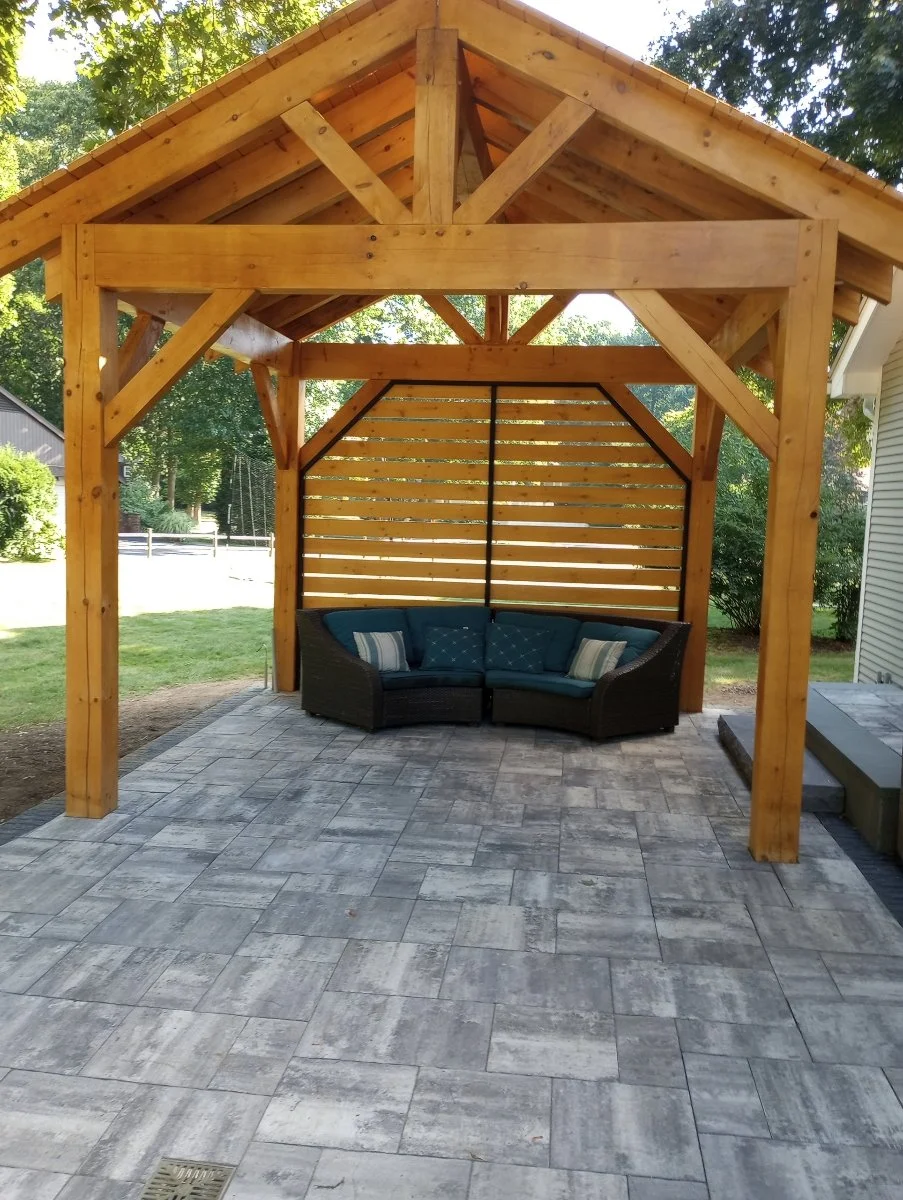 A backyard patio with a wooden pergola structure, and a corner wicker sofa with blue and striped cushions, on grey stone tiles, surrounded by greenery and trees.