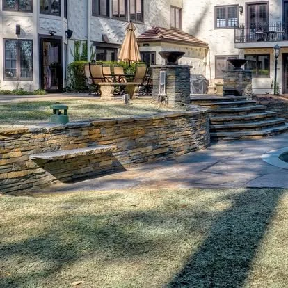 Backyard with stone patio, stairs, fountain, outdoor seating area with umbrella, and residential buildings in the background.