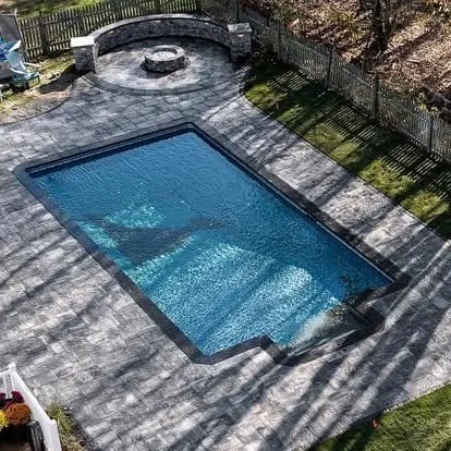 An outdoor swimming pool with a concrete patio surrounded by a wooden fence and some trees, with shadows cast across the scene.