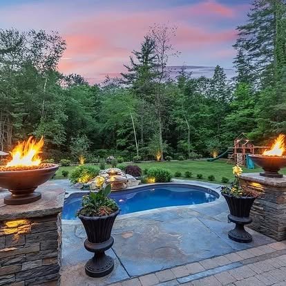 Backyard with a hot tub surrounded by stone deck, two fire bowls, and plants, with trees and a colorful sunset sky in the background.
