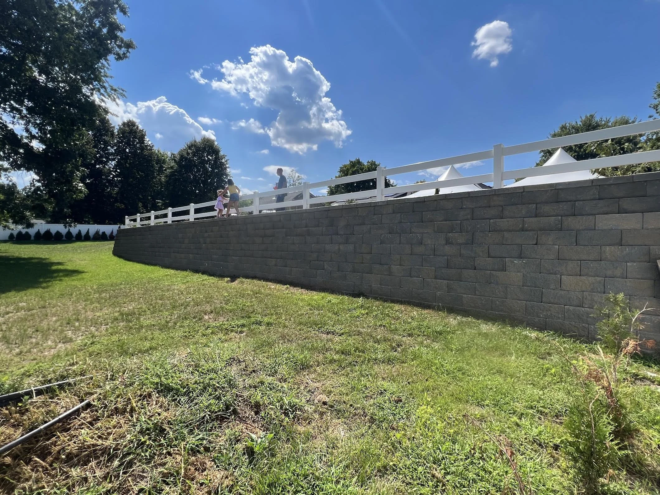 A grassy yard with a stone wall and white fence on top, with two children and an adult on the fence, and white tents in the background on a sunny day with blue sky and clouds.