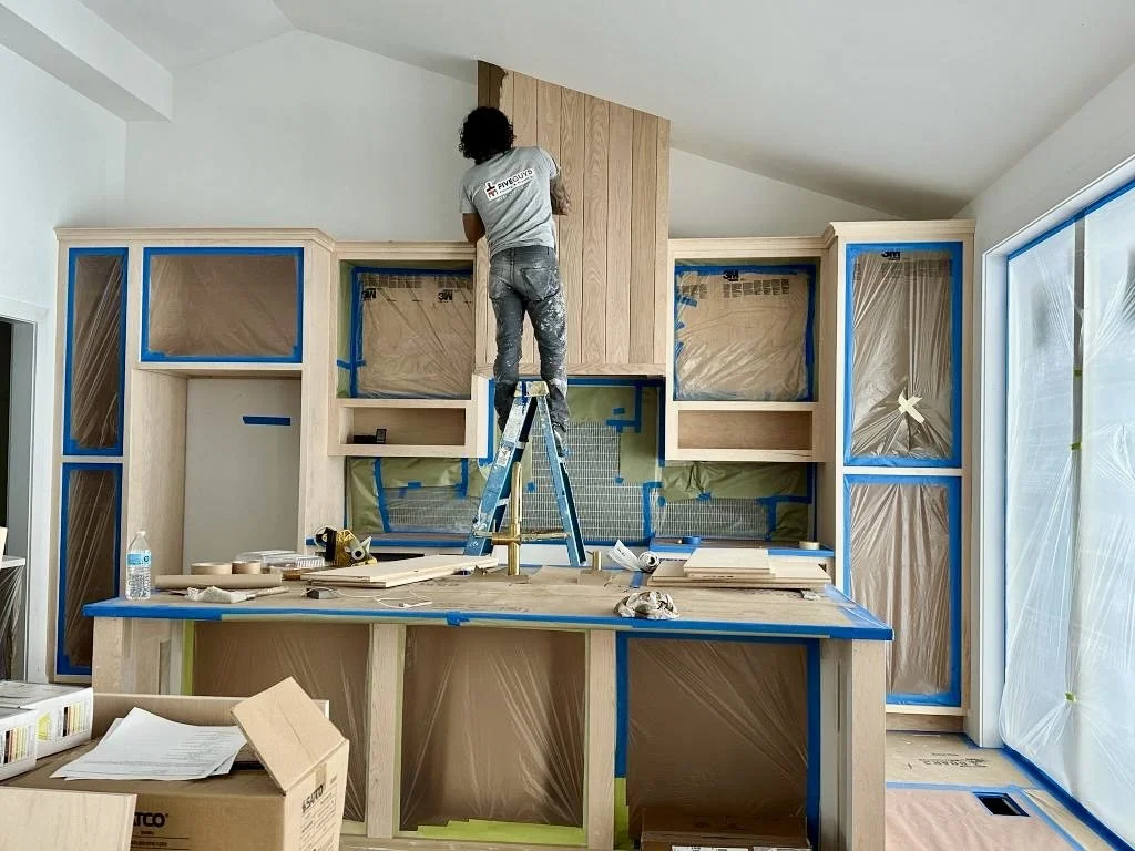 Cabinet painting prep with masked kitchen cabinets, protected surfaces, and painter on ladder during cabinet refinishing project.