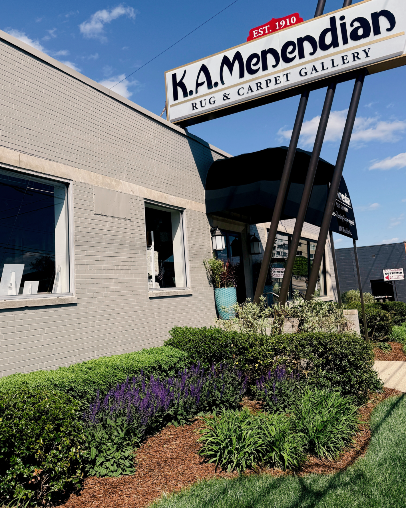 Exterior view of K. A. Mendendian Rug & Carpet Gallery store with a large signboard, black awning, flower pots, green bushes, and landscaped garden under a blue sky with some clouds.