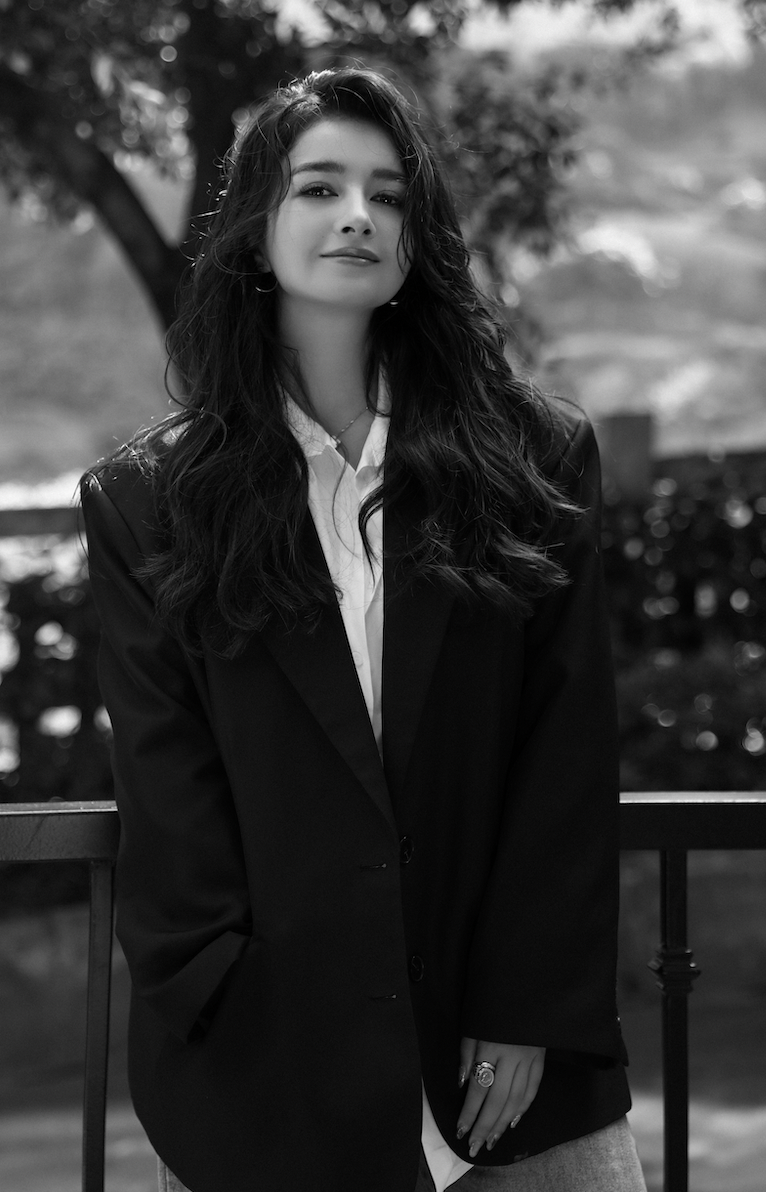 Black-and-white photo of a young woman with long, wavy hair, wearing a blazer over a shirt, standing outdoors next to a railing with a blurred natural background.