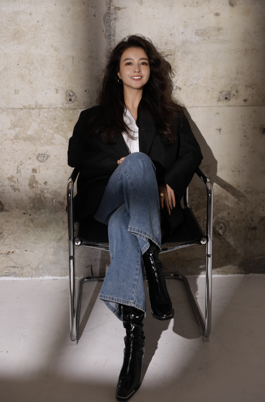 A young woman with dark hair sitting on a black chair against a concrete wall, wearing a black jacket, white top, blue jeans, and black shiny boots, smiling at the camera.
