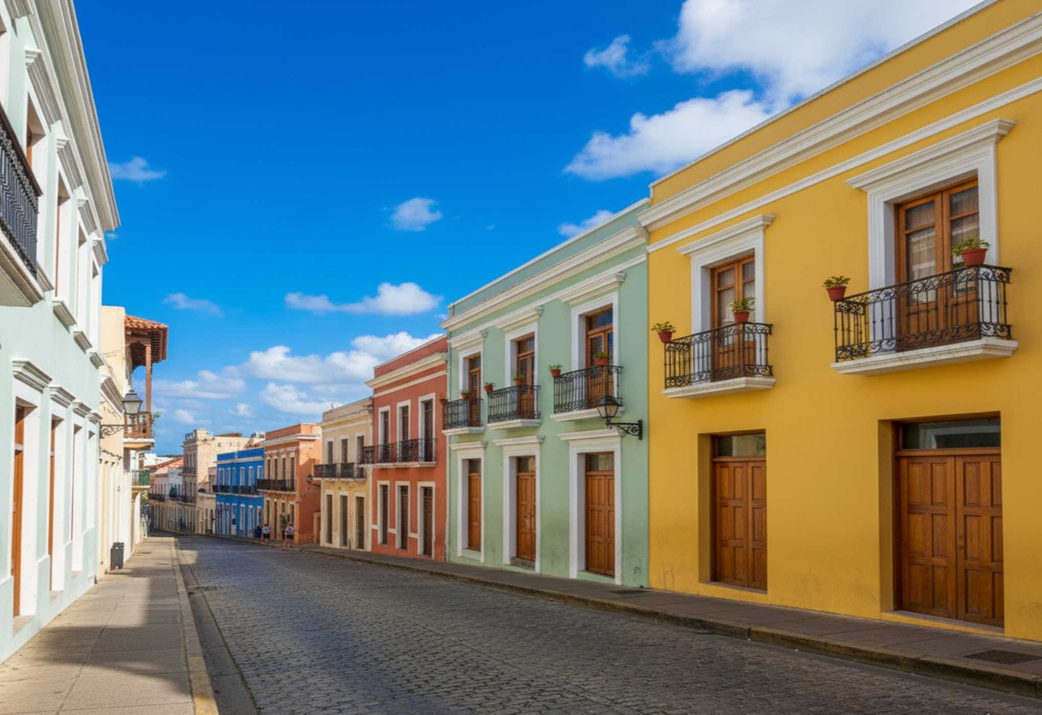Colorful colonial-style buildings with balconies and potted plants on a cobblestone street under a partly cloudy blue sky.