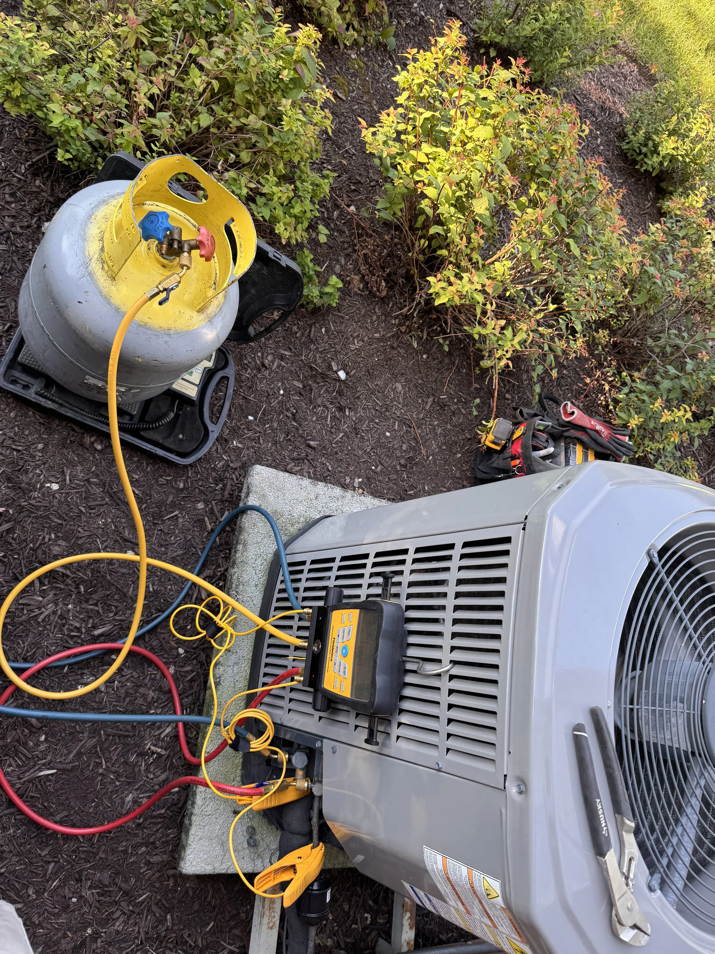 A portable air conditioning unit being serviced outdoors, with refrigeration gauges attached, a tank of refrigerant, and tools nearby, surrounded by bushes and mulch.