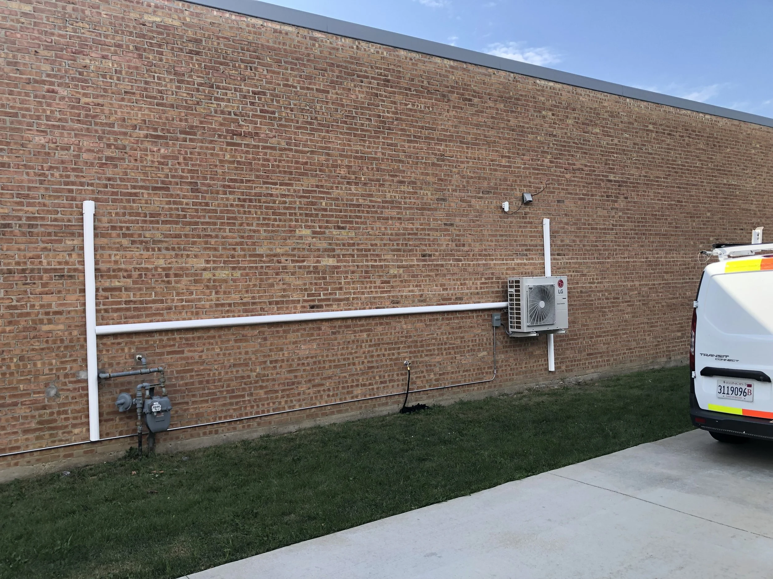 Exterior brick wall with HVAC system, pipes, utility meter, and a white utility van parked nearby.