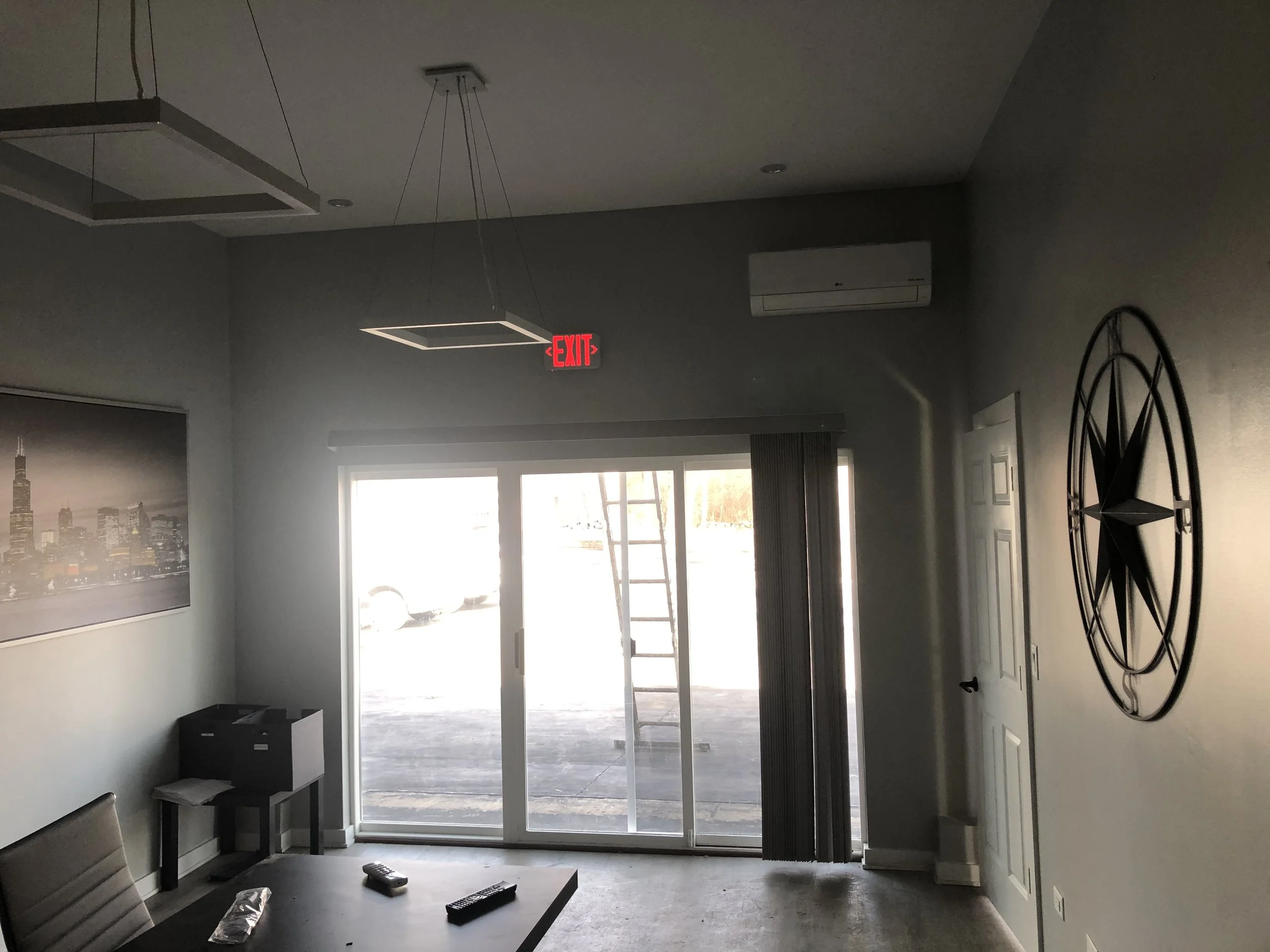 An empty room with a sliding glass door, a ladder outside, a chalkboard clock on the wall, a framed cityscape picture, a black side table, and a TV remote on a table.