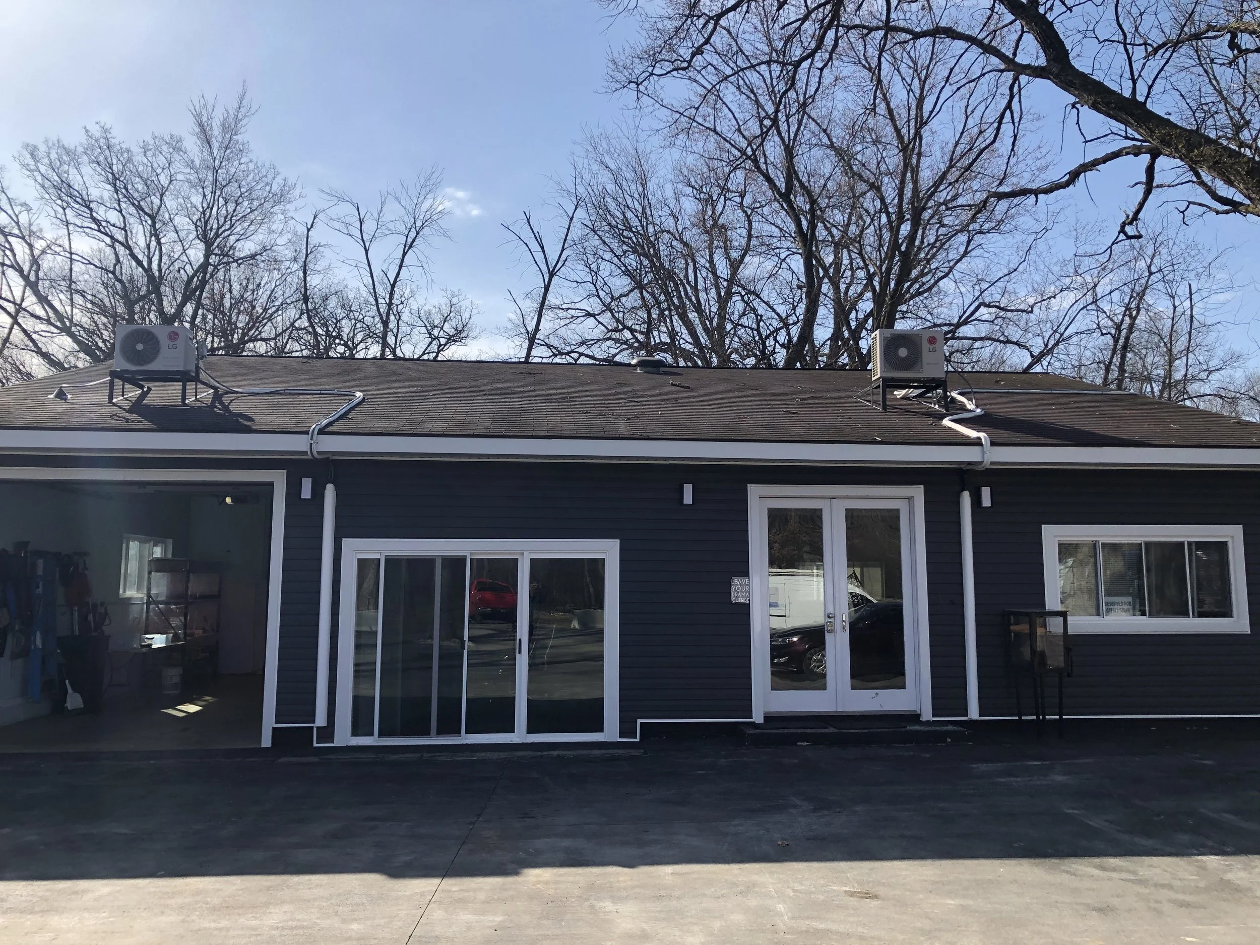 The image shows a black building with large windows and glass doors, with two air conditioning units on the roof and leafless trees in the background under a blue sky.