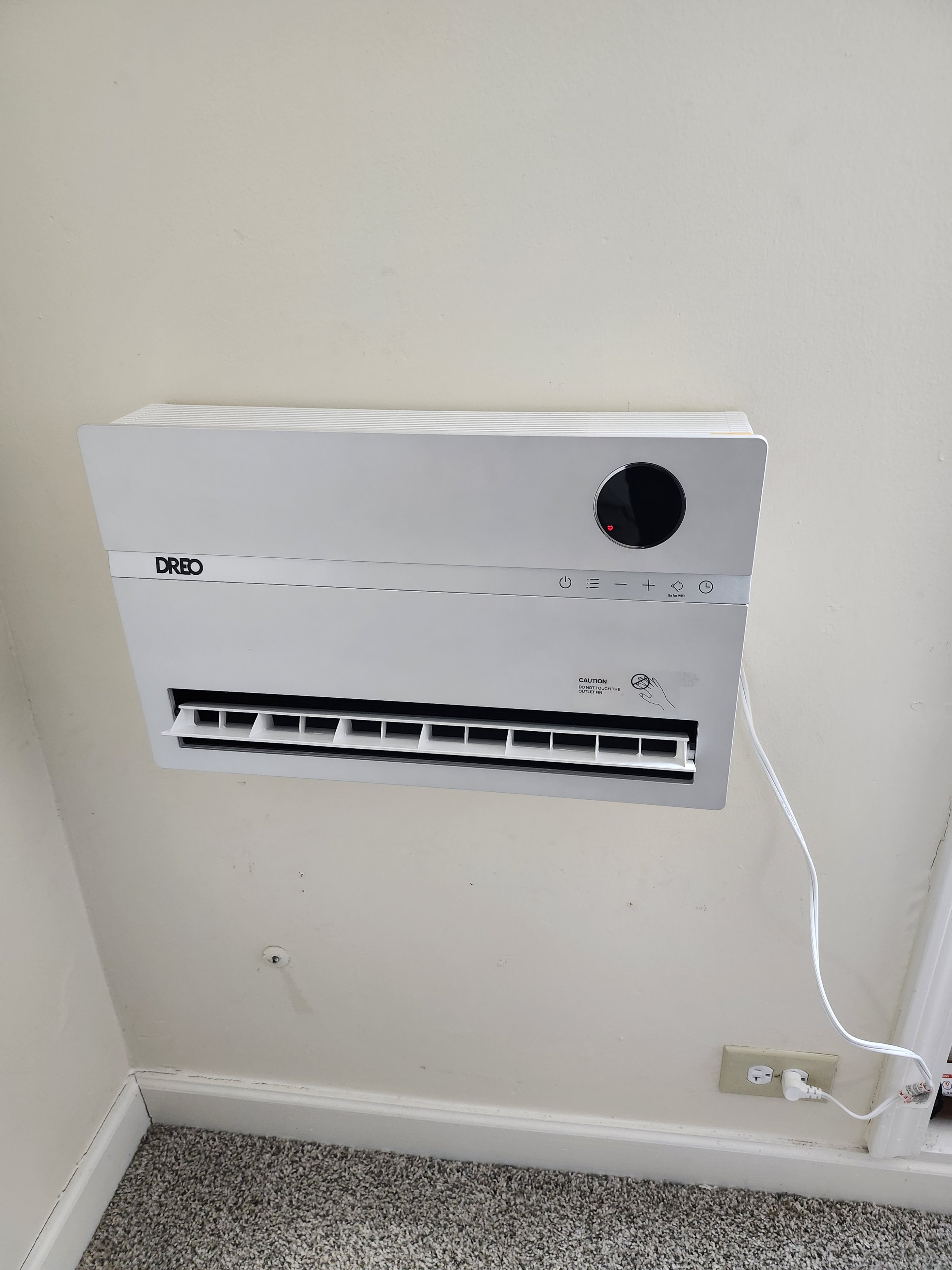 Wall-mounted white Dreo air conditioner with a digital display and control buttons, plugged into a wall outlet, above a carpeted floor.
