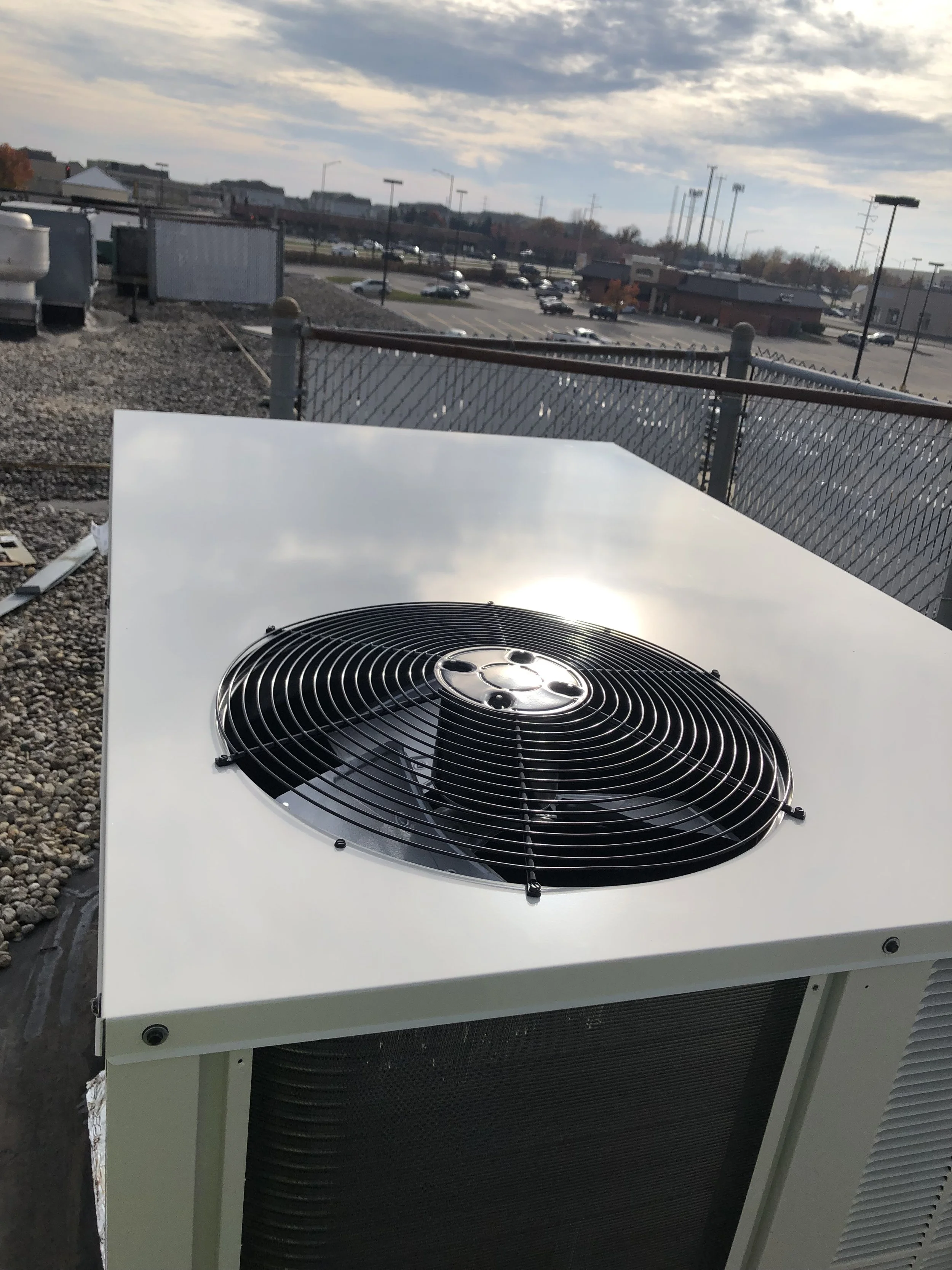 A rooftop air conditioning unit with a fan and grille, overlooking a parking lot and commercial buildings in the distance under a partly cloudy sky.