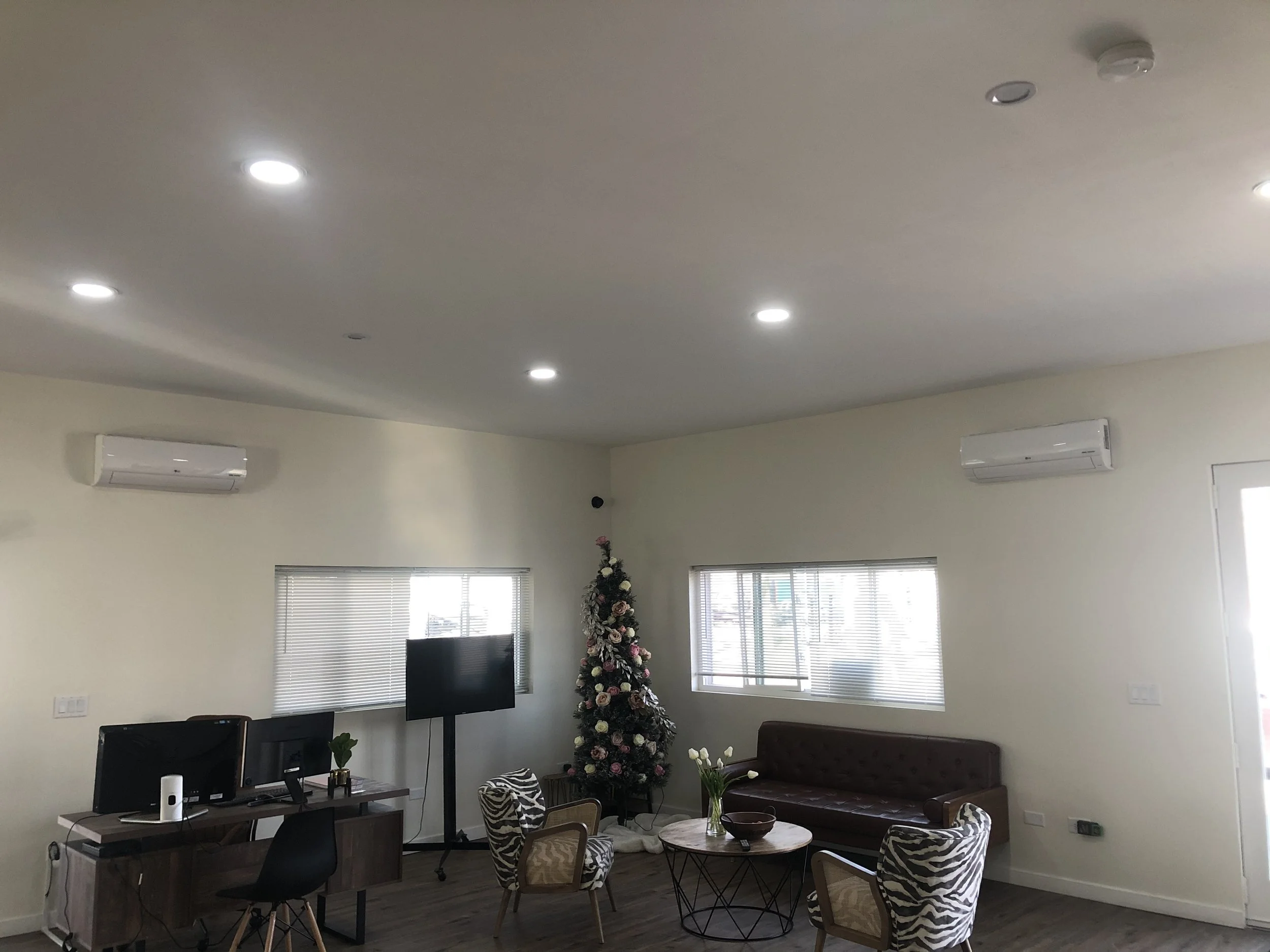 Living room with Christmas tree, flat-screen TV, two air conditioners, large windows with blinds, sofa, round coffee table with flowers, two zebra-patterned armchairs, and wooden flooring.