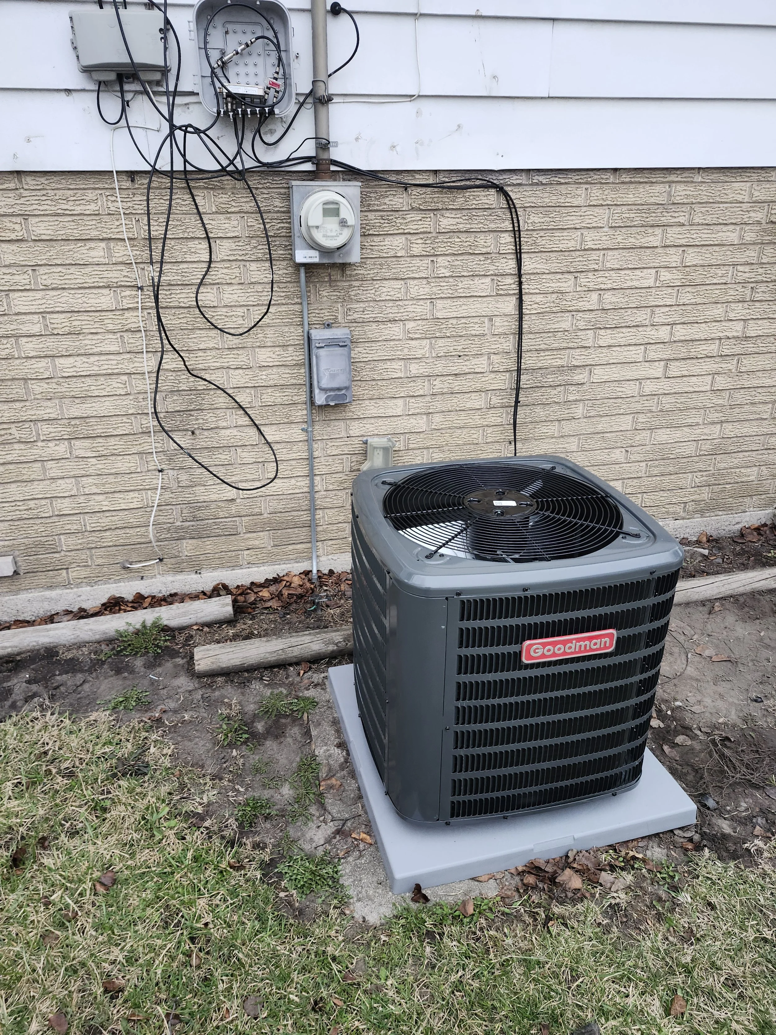 Outdoor view of a Goodman air conditioning unit installed on a concrete pad next to a house wall. The background includes electrical meters, conduits, and wiring on the house's exterior wall.