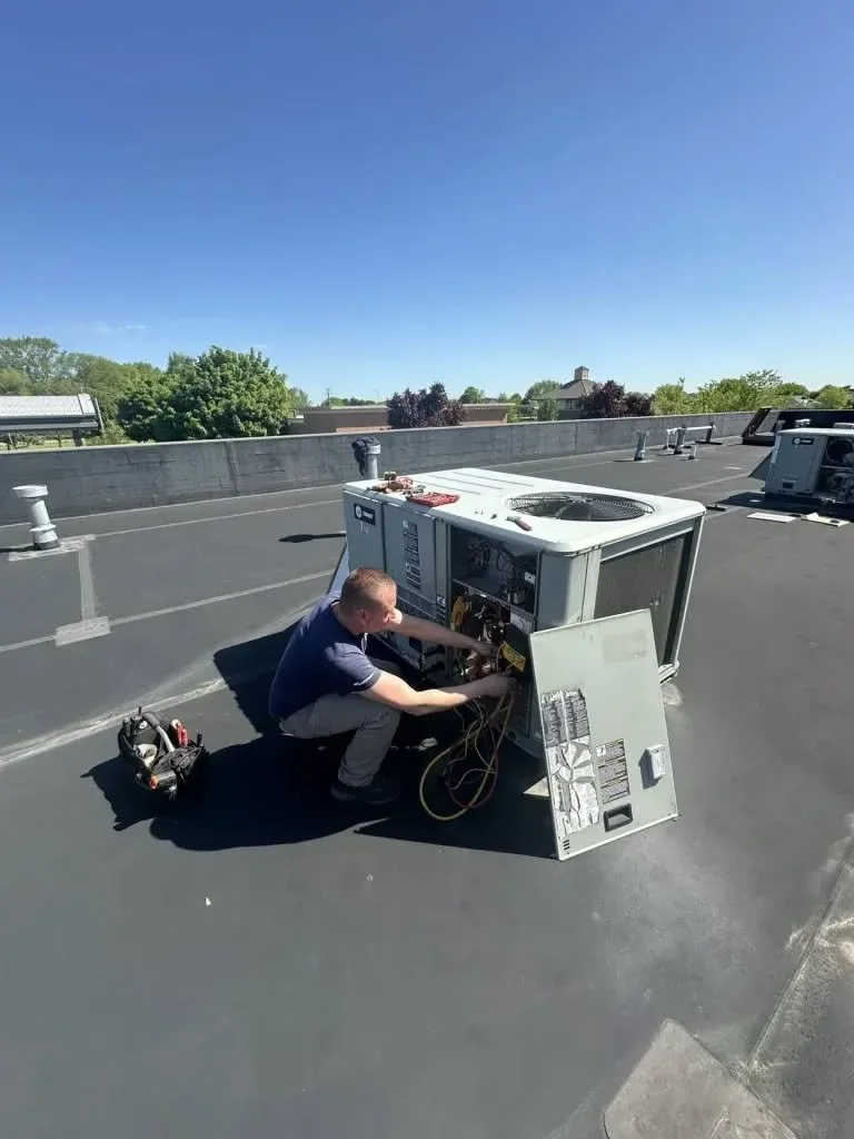 A person working on repairing or maintaining air conditioning or HVAC equipment on a flat rooftop under a clear blue sky.