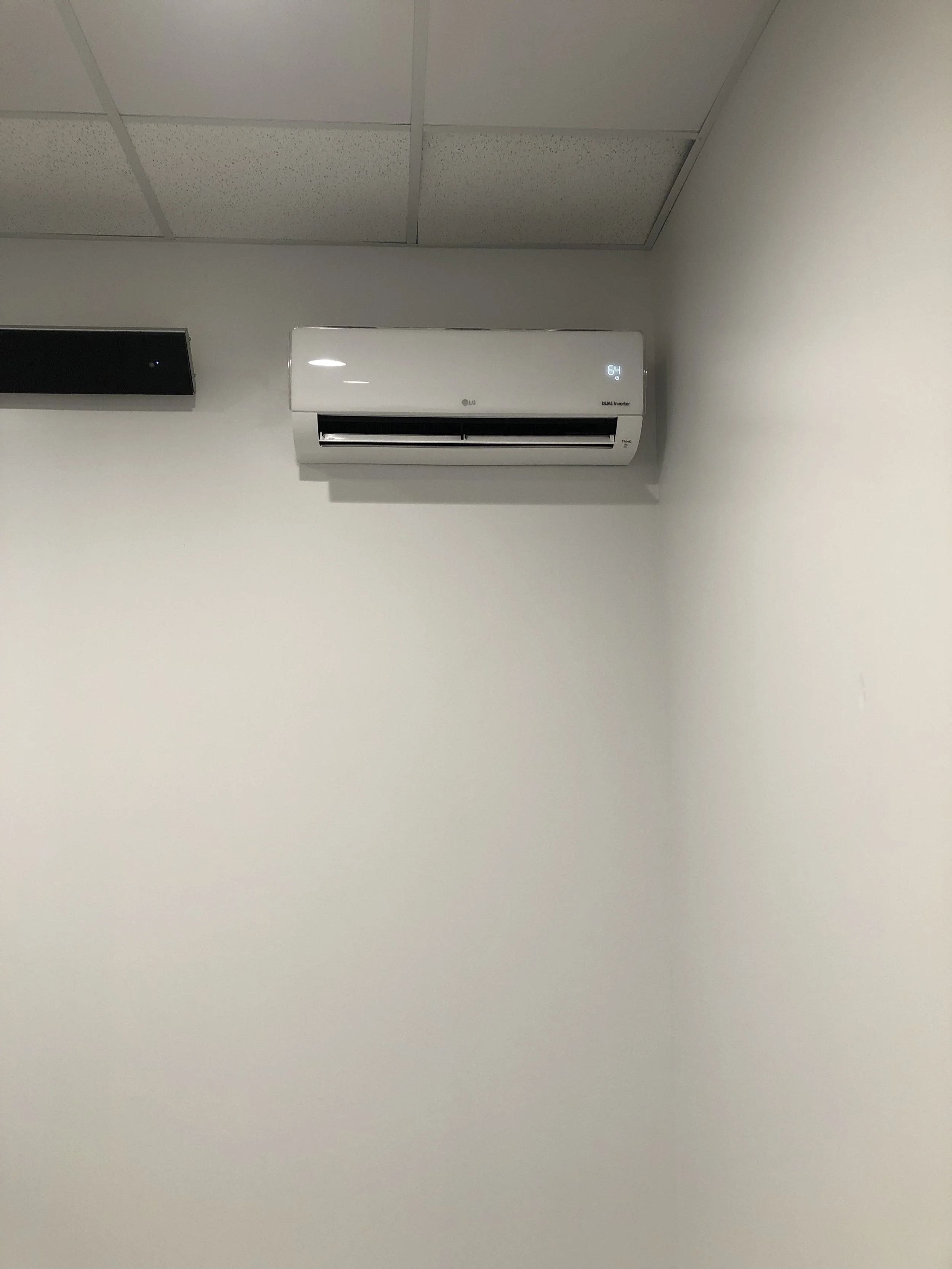 Wall-mounted air conditioner unit with digital display reading 64 degrees, installed on a plain white wall in an office or room.