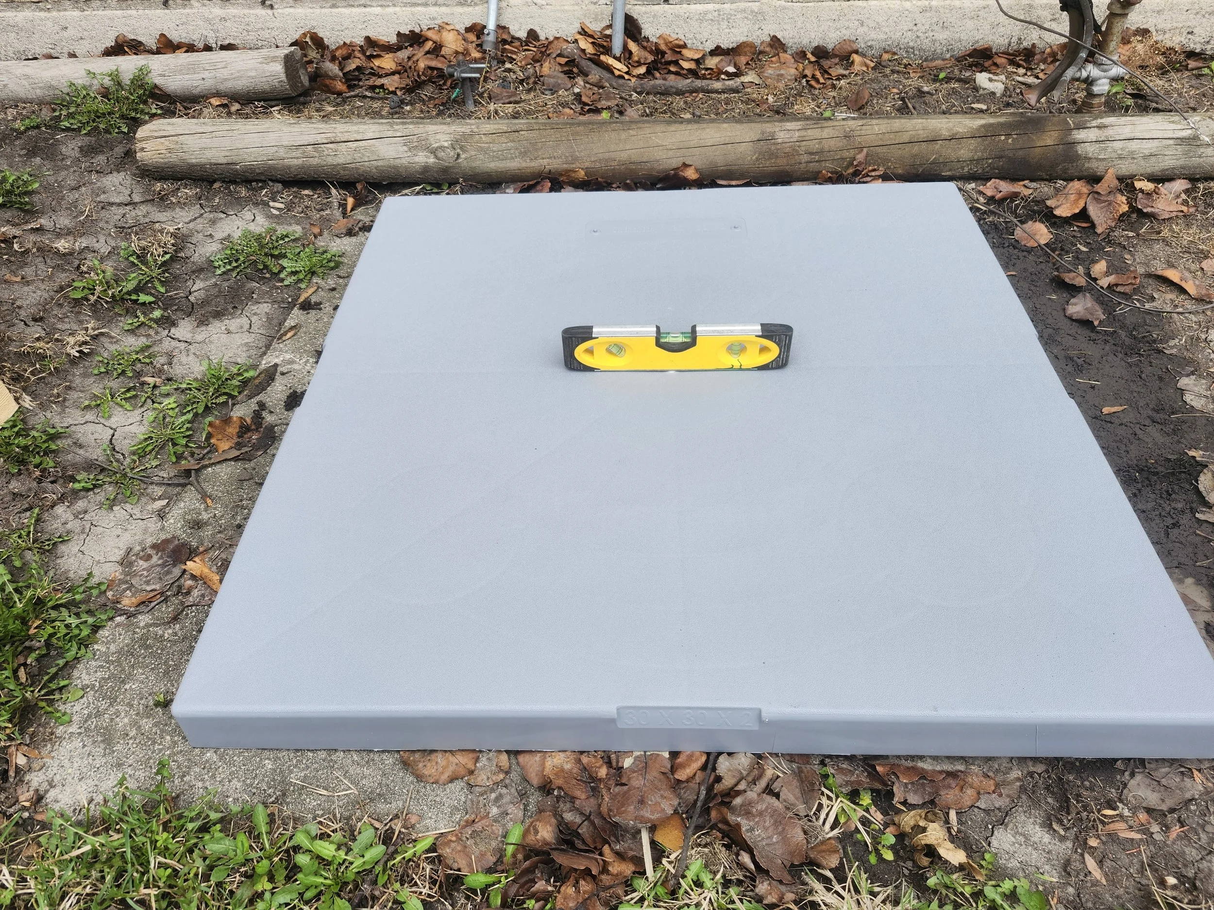 A gray access panel cover with a built-in level tool on top, placed on outdoor soil with scattered leaves, small green plants, and some wooden logs and electrical wires in the background.