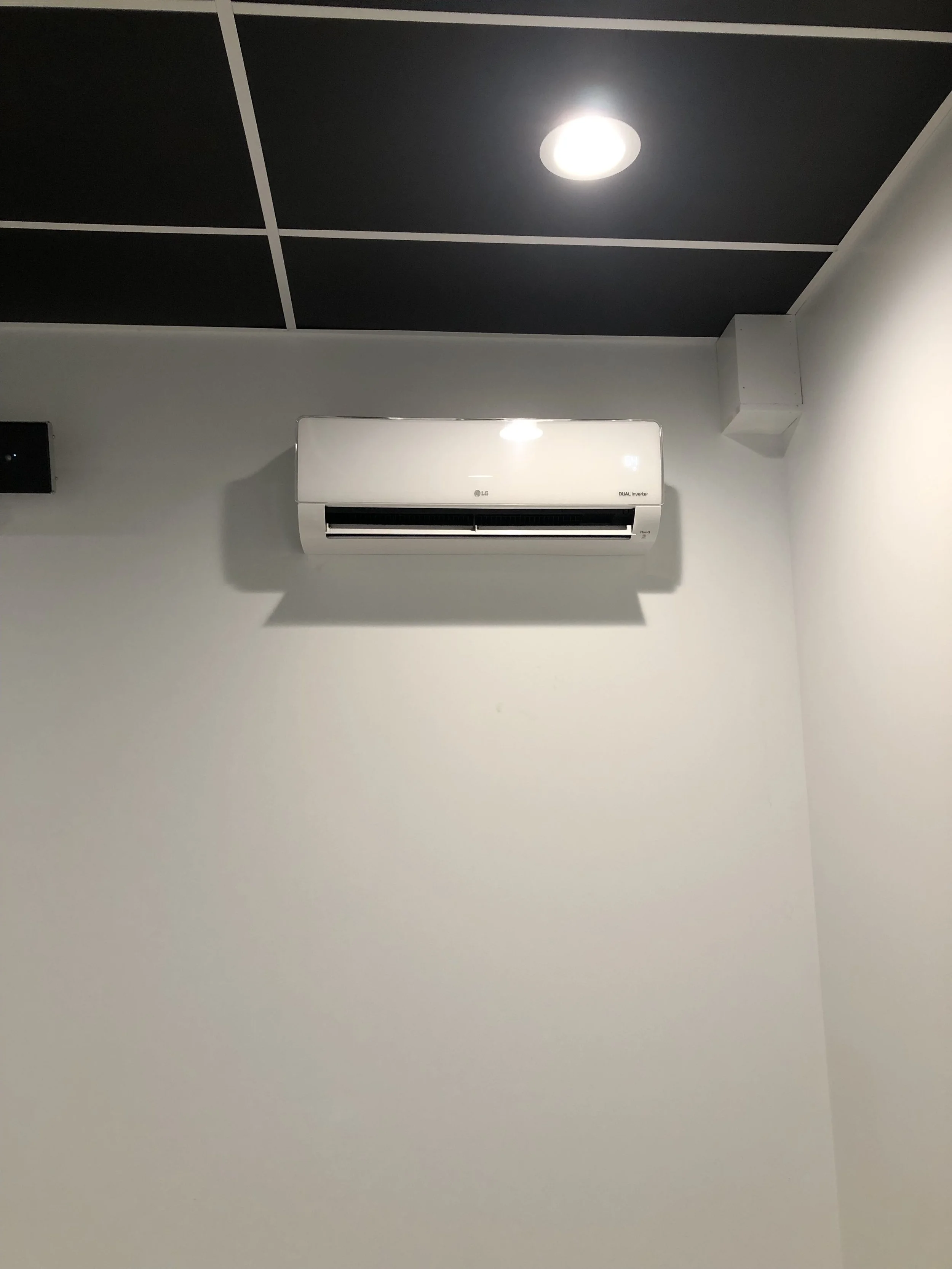 White wall-mounted air conditioner unit with black vents in a room with black ceiling tiles and a ceiling light.