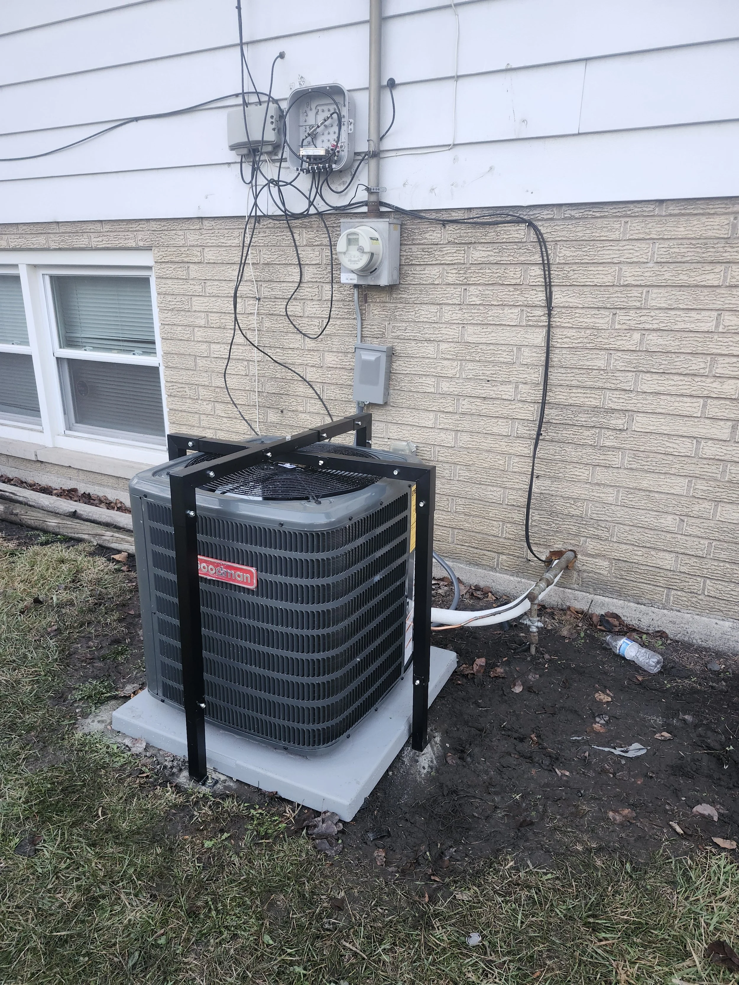 Outdoor air conditioning unit with black protective frame mounted on a concrete pad outside a house, with electrical wiring and meters on the wall behind.