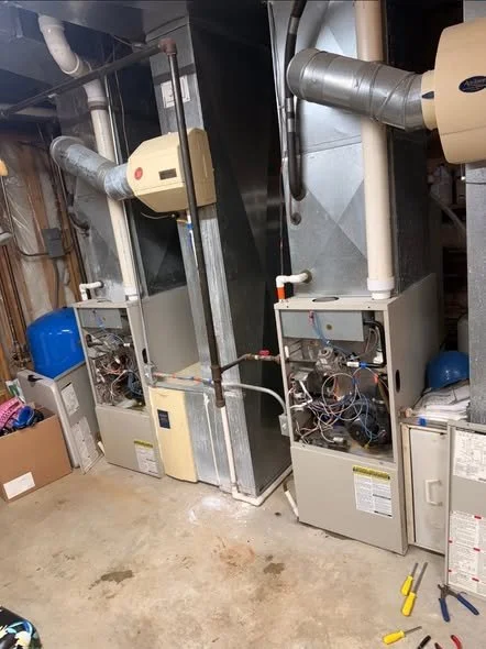 Two gas furnaces with visible internal components and ductwork in a basement utility room, with tools on the floor nearby.