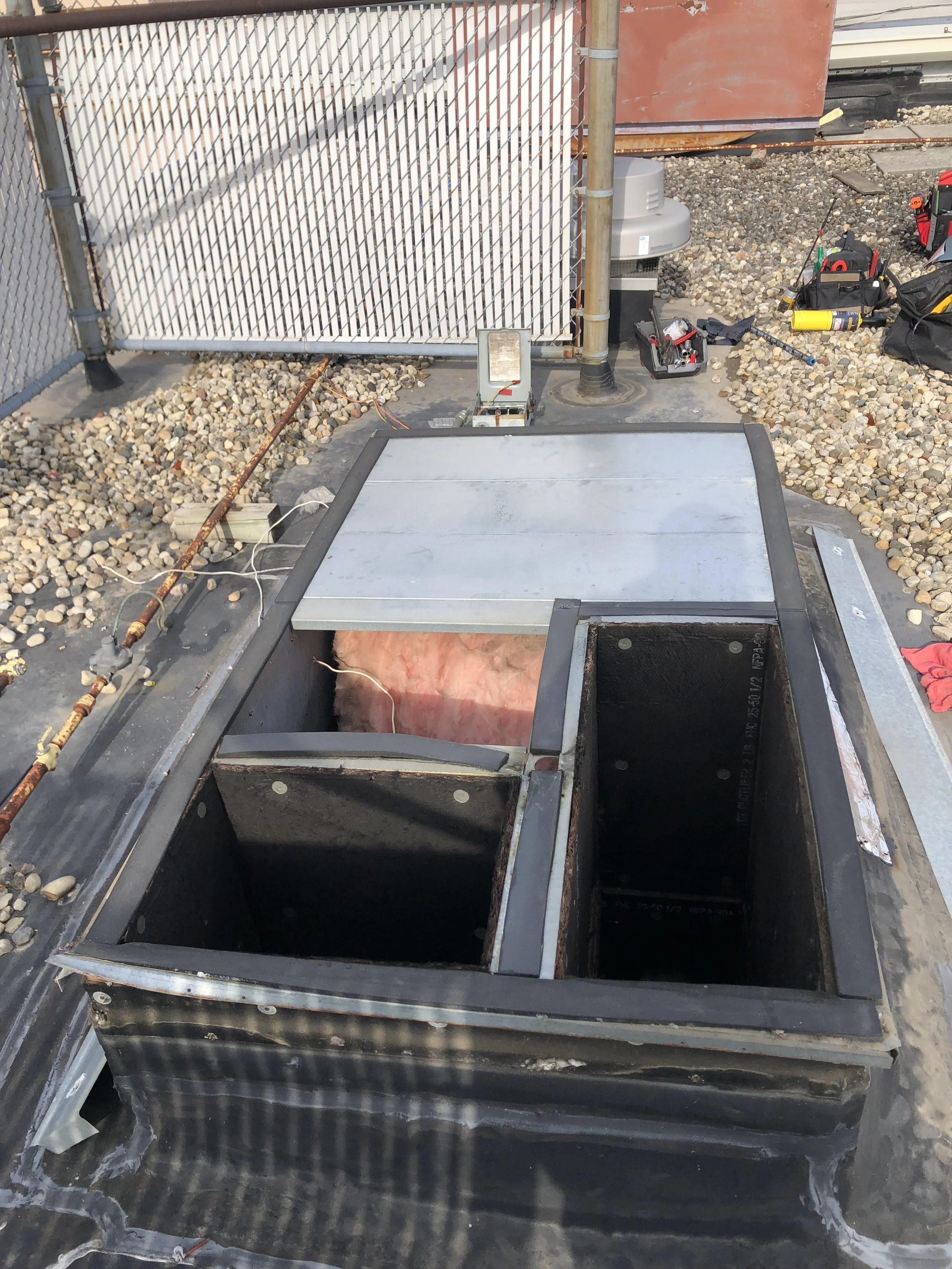 Open access hatch on a rooftop with insulation and construction tools around, surrounded by gravel and a metal fence.
