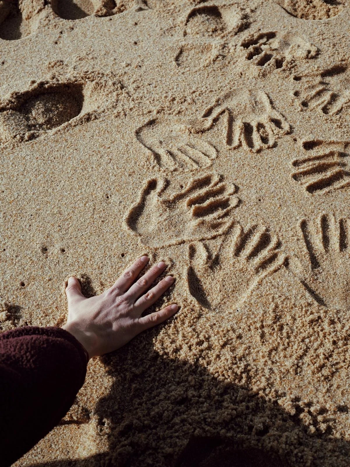 A person's hand touching sand with a starfish drawing and various footprints in the sand.
