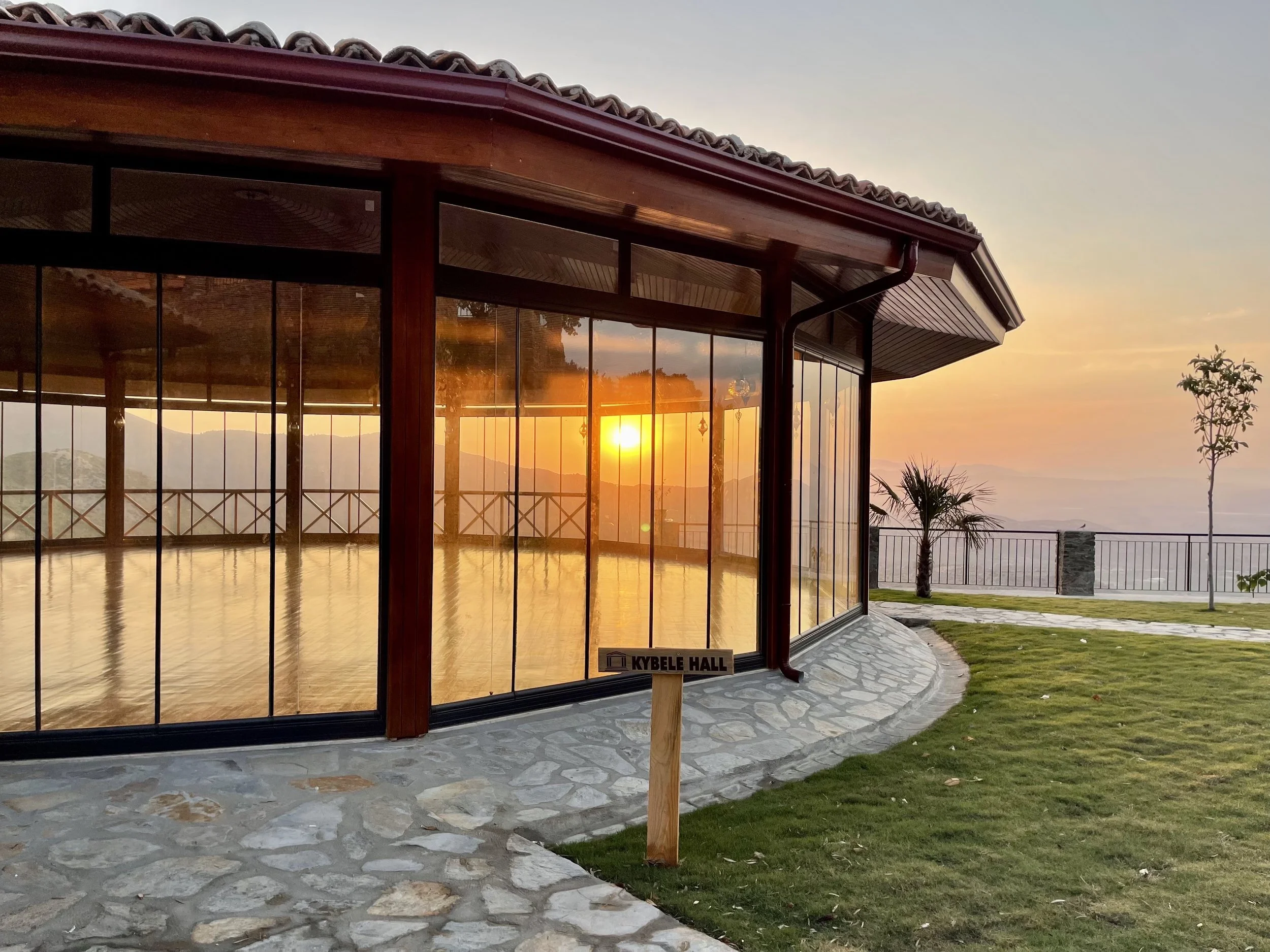 A glass-enclosed room with wooden framing reflects a sunset overlooking the mountains, with a stone patio and well-maintained grassy area outside, featuring a small sign reading 'KYEBLE HALL'.
