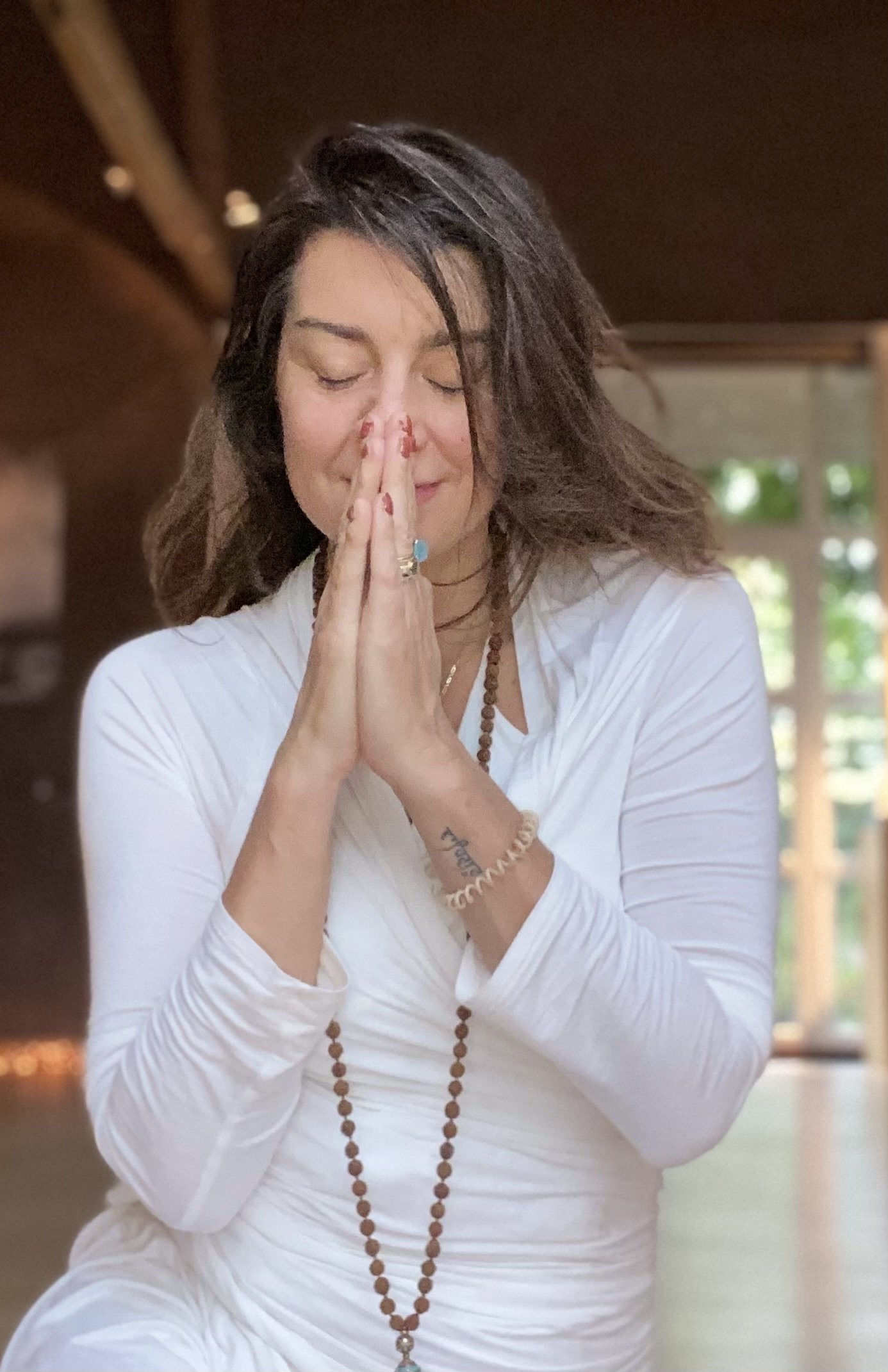 A woman with closed eyes, hands pressed together in a prayer or meditative position, wearing a white long-sleeve shirt, a beaded necklace, and rings, standing indoors with natural light in the background.
