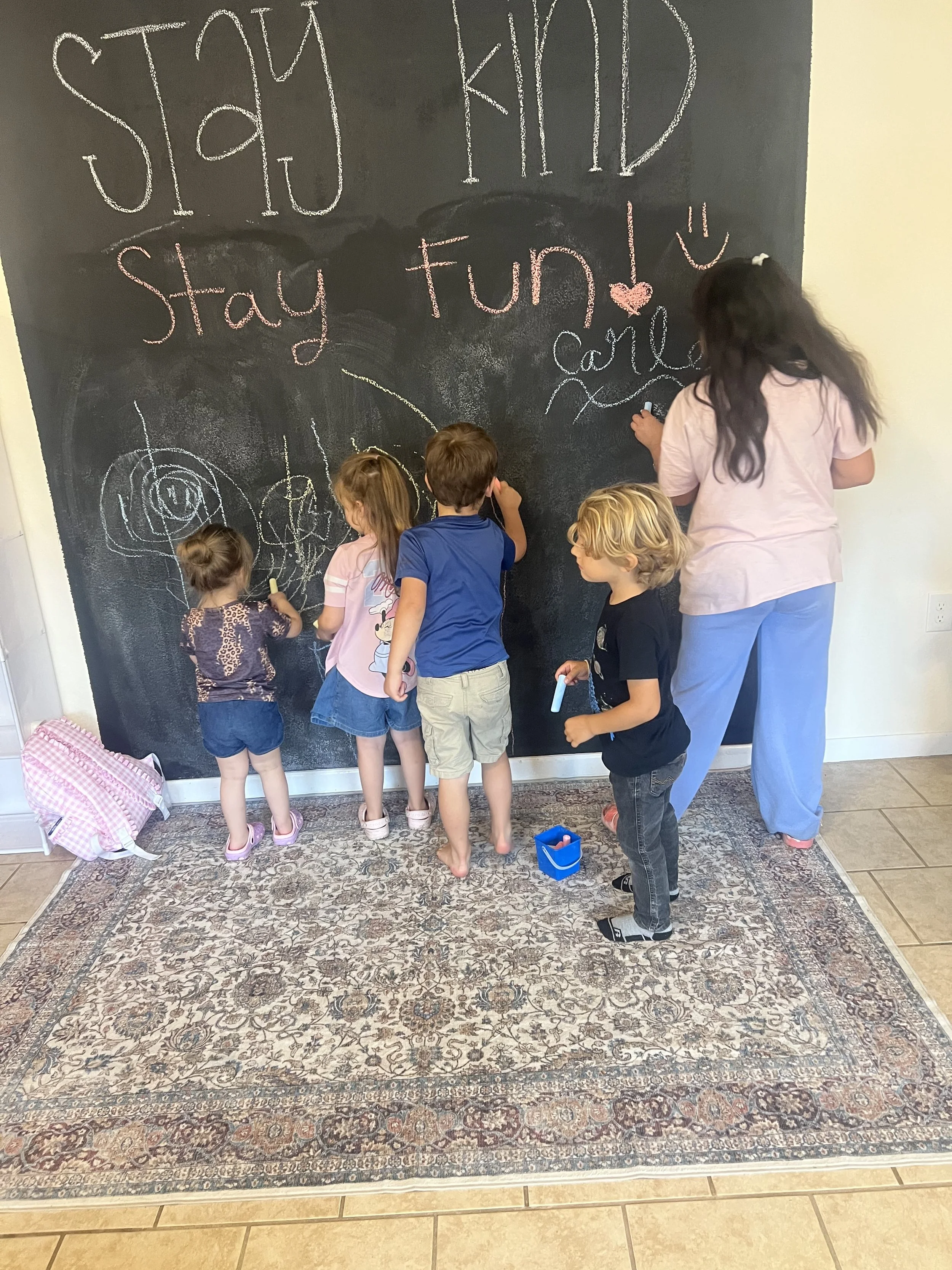 Children and an adult girl draw on a large black chalkboard wall with colorful chalks. The wall has a message written in different colors that says, "Stay KID! Stay fun!" The children appear engaged and creative.