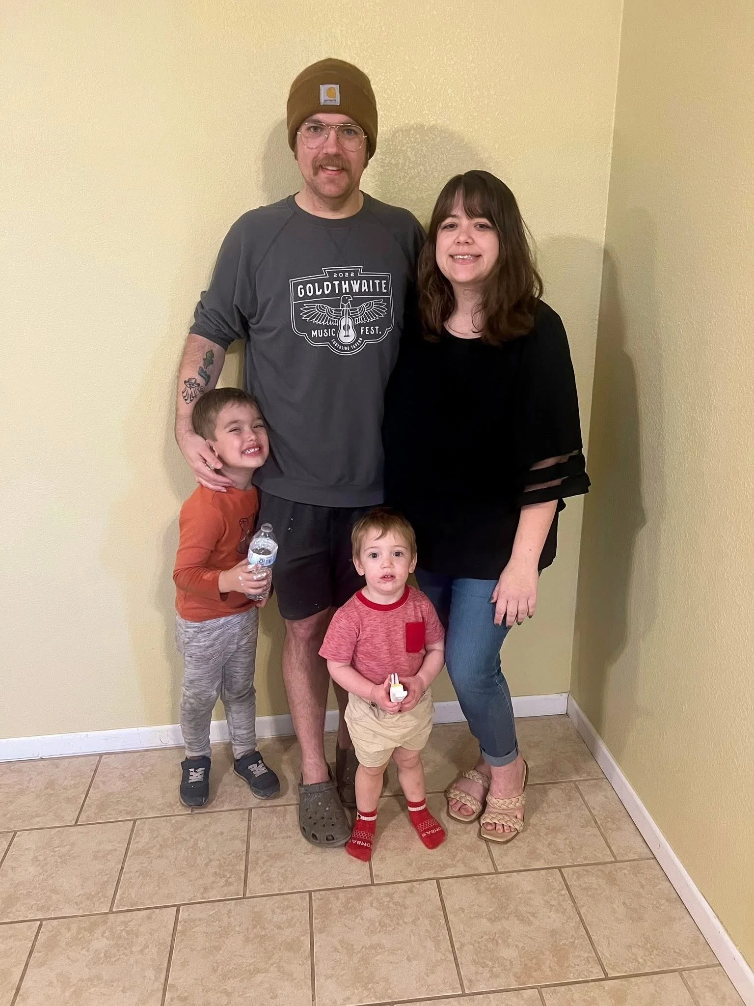 A family of four standing together indoors against a yellow wall, smiling at the camera. The man is wearing a brown beanie, glasses, a dark gray T-shirt, and shorts. The woman is dressed in a black top and jeans. The two young boys are standing in front; one is holding a water bottle and the other holds a small toy.