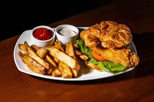 Fried chicken sandwich with lettuce, tomato, and fried chicken fillet on a bun, served with French fries, ketchup, and ranch dressing on a white plate.