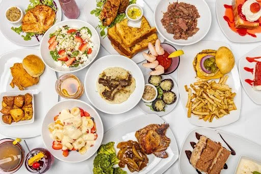 A variety of American comfort foods on white plates, including fried chicken, tacos, burger sliders, French fries, macaroni and cheese, steak, mashed potatoes, and desserts, on a white table.
