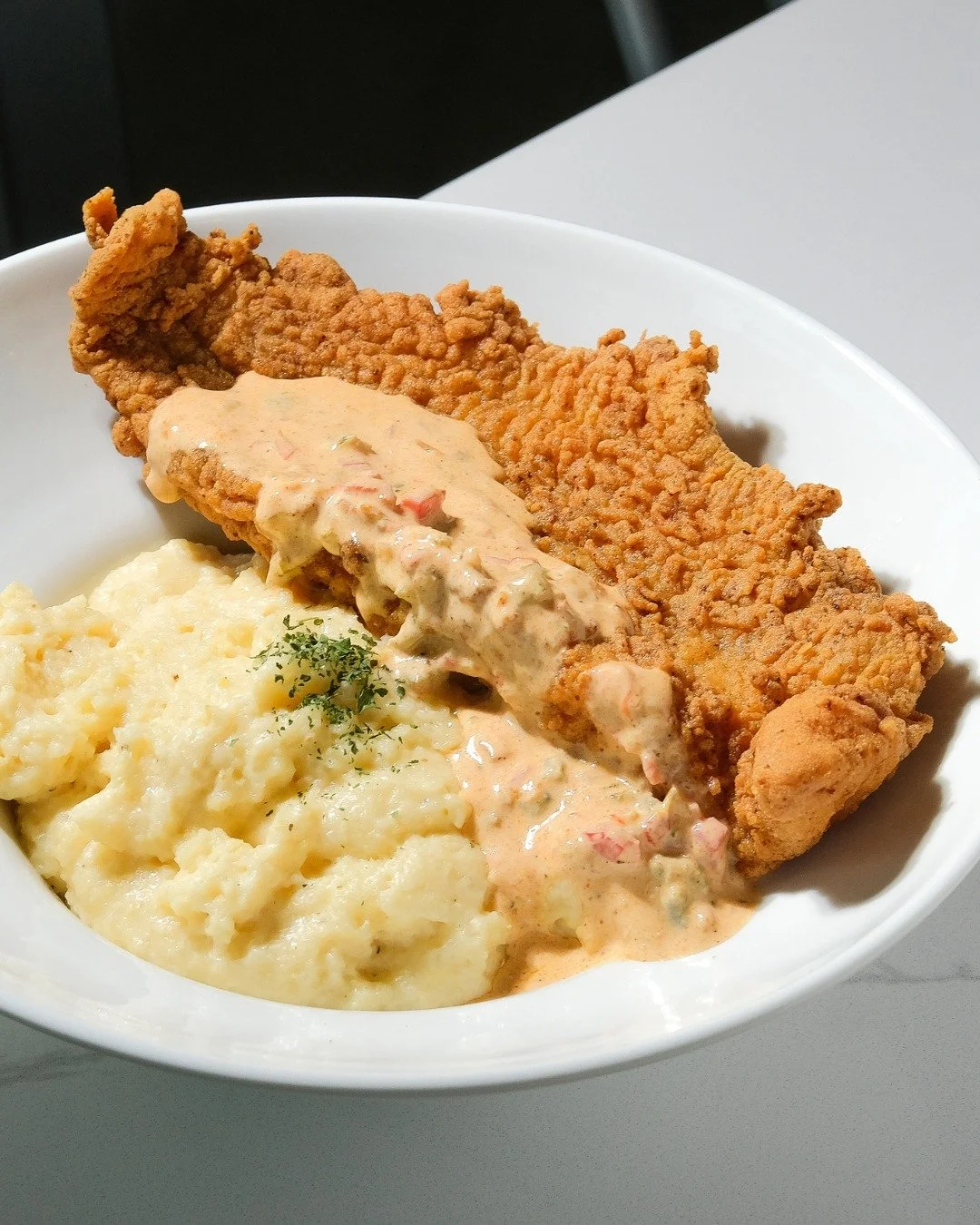 Fried chicken fillet with gravy, mashed potatoes with parsley, and a piece of fried fish in a white bowl.
