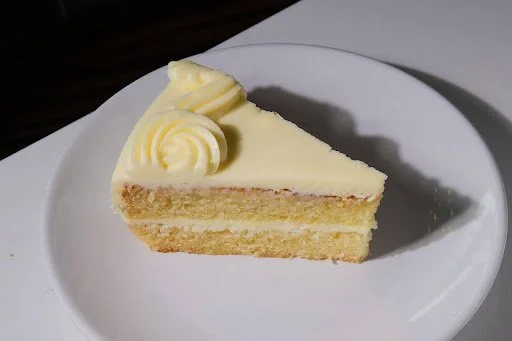 A slice of vanilla cake with white frosting and decorative cream on top, served on a white plate.