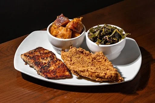 A plate with fried fish, a scoop of cooked sweet potatoes, a bowl of greens, and a fried chicken breast.