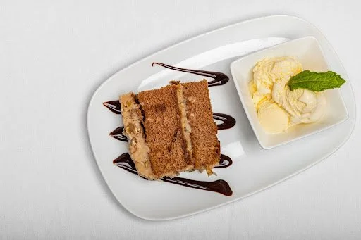 A slice of layered cake with chocolate drizzle on a white plate, served with two scoops of vanilla ice cream garnished with a mint leaf.