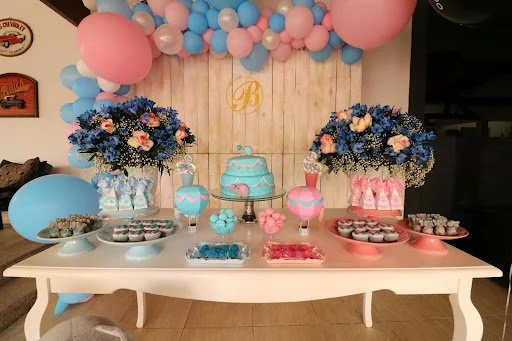 Decorative dessert table with blue and pink themed cupcakes, cake, and candy, adorned with blue and pink balloons and floral arrangements for a celebration.