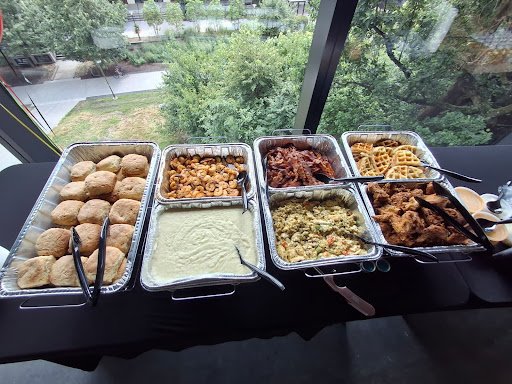 Buffet table with various dishes including chicken nuggets, macaroni and cheese, barbecue ribs, waffles, mashed potatoes with gravy, and fried chicken, set next to a large window with a view of trees and a street.