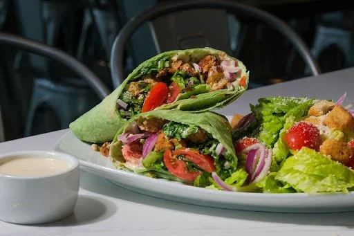 A plate with a large wrap burrito and a side salad, with a small bowl of white sauce on a white table in a restaurant setting.