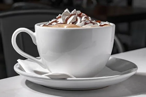 A white mug filled with coffee topped with whipped cream and drizzled with chocolate, placed on a white saucer with a spoon.