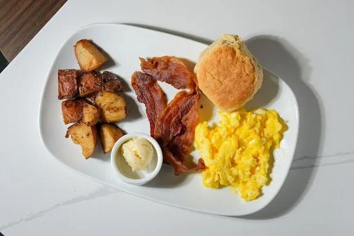Breakfast plate with home fries, bacon, a biscuit, scrambled eggs, and a small cup of butter.