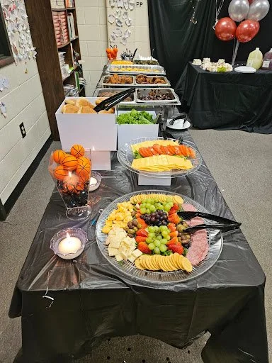 A buffet table with fruit platter, cheese, grapes, and various food items, decorated with orange and silver balloons, candles, and a black tablecloth in a room.